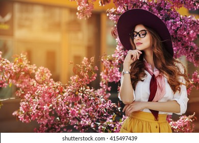 Outdoor portrait of young beautiful fashionable lady posing near flowering tree. Model wearing stylish accessories & clothes. Girl closed her eyes. Female beauty & fashion. City lifestyle. Copy space 