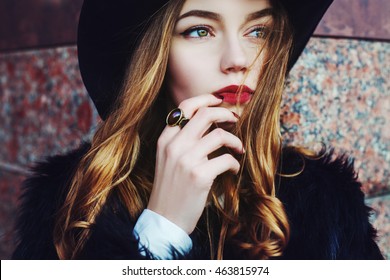 Outdoor portrait of a young beautiful fashionable lady wearing stylish black fur coat and wide-brimmed hat. Model looking aside. Female fashion concept. City lifestyle. Close up.