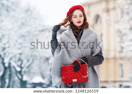 Outdoor portrait of young beautiful  fashionable woman wearing stylish winter gray coat, woolen scarf, red beret, leather gloves, holding quilted bag, posing in street of european city. Copy space
 