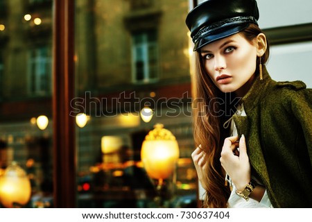 Outdoor portrait of young beautiful fashionable woman posing in street. Model wearing stylish leather hat, green jacket. Lights and reflections on background. Copy, empty space for text