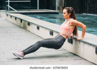 Outdoor portrait of young beautiful fit woman doing tricep dips, wearing fashionable activewear, athlete model posing in urban background, sport fashion