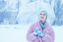 Outdoor Portrait Of Young Beautiful Happy Smiling Girl Walking In Snow-covered Street. Model Wearing Trendy Winter Pink Faux Fur Coat, Blue Beanie Hat, Gloves, Holding Transparent Umbrella. Copy Space