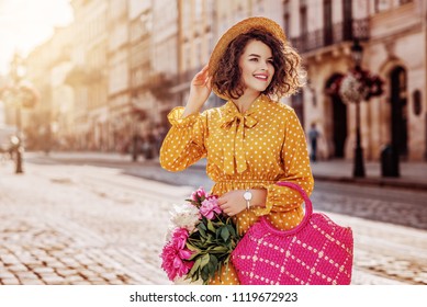 Outdoor Portrait Of Young Beautiful Happy Smiling Girl Wearing Yellow Polka Dot Dress, Hat, Holding Straw Bag With Peonies, Posing In Street Of European City. Spring, Summer Fashion. Copy Space 