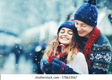  Outdoor portrait. Young beautiful happy smiling couple posing on street.  Models hugging, looking at candy canes, wearing stylish winter clothes. Snowfall. City lifestyle. Copy, empty space for text