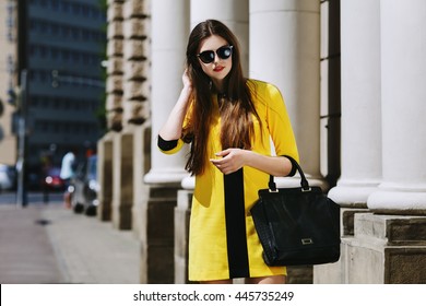 Outdoor Portrait Of Young Beautiful Lady Walking On The Street. Model Wearing Sunglasses & Stylish Yellow Summer Dress. Girl Looking Down. Female Fashion Concept. City Lifestyle. Sunny Day. Waist Up