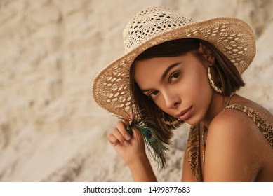 Outdoor portrait of young beautiful woman with luxury tanned skin wearing straw hat, snakeskin print swimwear, hoop earrings, posing in sand desert, at sunset. Copy, empty space for text 