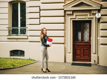 Outdoor portrait of young beautiful woman holding white box with red roses, wearing leather jacket and checked trousers