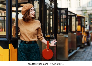 Outdoor Portrait Of Young Fashionable Woman Wearing Beige Beret, Turtleneck, Green Corduroy Trousers, Belt, Holding Round Orange Suede Handbag, Posing In Street Of European City. Copy Space For Text