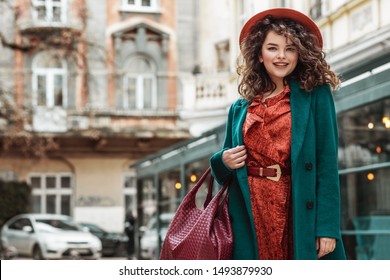 Outdoor Portrait Of Young Happy Smiling Fashionable Lady Wearing Trendy Orange Dress, Hat, Belt, Green Classic Coat, Holding Red Handbag, Posing In Street Of European City. Copy, Empty Space For Text