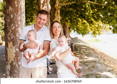 Outdoor portrait of young happy smiling mother and father with twin babies on natural background with boats and maple trees. Happy family wearing white clothes holding baby boy and baby girl twins.