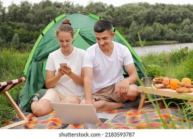 Outdoor Portrait Of Young Pretty Hiker Couple Sitting Near A Tent Looking At Laptop In The Nature, Woman Using Cell Phone, Family Spending Time In Open Air, Enjoying Resting.