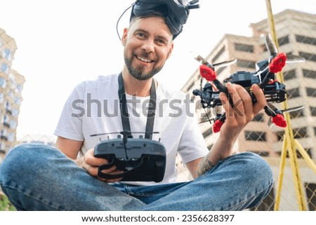 Outdoor portrait of young smiling fpv drone pilot wearing goggles and posing with remote controller and copter in hands. Happy winner celebrating victory in multicopter racing competition.