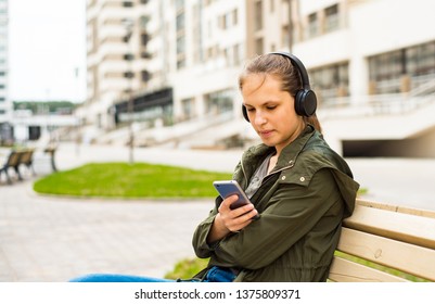 Outdoor portrait of young teenager brunette girl with long hair. young woman listening to music using mobile phone sitting on bench - Shutterstock ID 1375809371