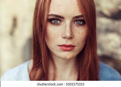 Outdoor portrait of young tender redhead student girl with healthy freckled skin in glasses looking at camera with natural expression on summer sunset