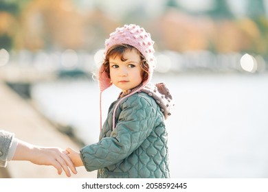 Outdoor portraot of cute toddler girl walking by the lake, holding mom's hand, looking back over the shoulder, wearing patch jacket and pink hat, cold weather