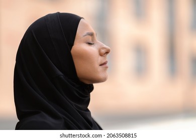 Outdoor Profile Portrait Of Calm Young Muslim Woman In Black Hijab Standing With Closed Eyes, Side View Shot Of Peaceful Religious Islamic Female Wearing Headscarf Standing On City Street, Closeup