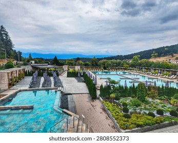 An outdoor resort spa complex features multiple swimming pools with lounge chairs, umbrellas, and landscaped gardens against a mountain backdrop. Photo taken in hotsprings in Bania, Bulgaria.