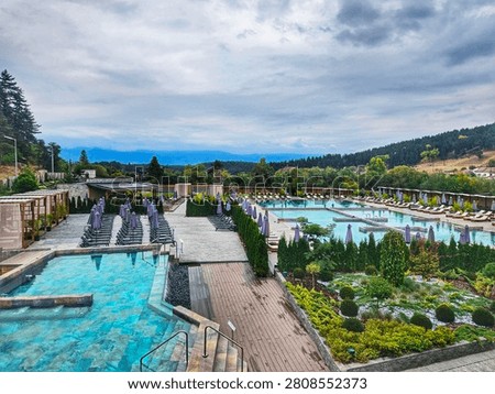 An outdoor resort spa complex features multiple swimming pools with lounge chairs, umbrellas, and landscaped gardens against a mountain backdrop. Photo taken in hotsprings in Bania, Bulgaria.