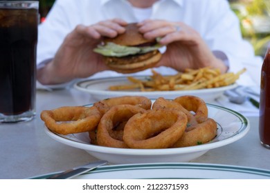 At An Outdoor Restaurant, A Plate Of Onion Rings Is On The Table Along With A Soda And Ketchup, And A Person Is Eating A Veggie Burger And Fries In The Background.