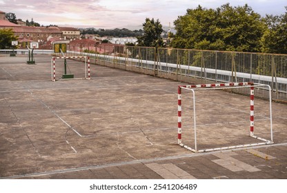 Outdoor rooftop sports court featuring worn, marked concrete ground with two basketball hoops and handball goals, surrounded by a high fence, set against urban rooftops - Powered by Shutterstock