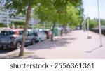 Outdoor scene in a sunny city with blurred cars parked along a street lined with trees and a sidewalk.