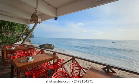 Outdoor seaside dining area with several red chairs and tables set up on a wooden deck. The view from the dining area includes a calm sea, clear sky, and a horizon line where the sky meets the water. - Powered by Shutterstock