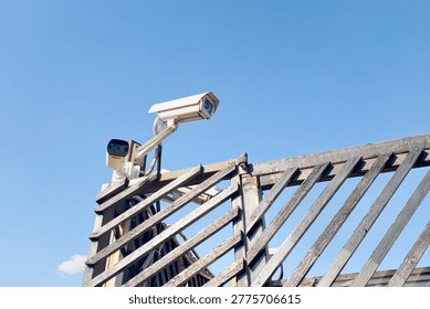 Outdoor Security Camera System Mounted On Wooden Structure. Two surveillance cameras installed on a wooden gate against blue sky. Property protection and perimeter security monitoring equipment