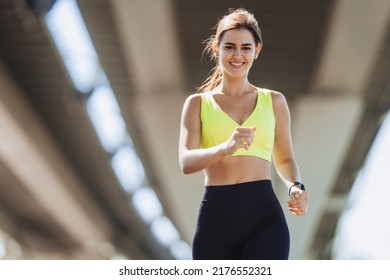 Outdoor Shoot Of Young Brunette Tanned Woman Running Under Overpass, Smiling Broadly Looking At Camera. Gorgeous Hispanic Girl In Yellow Bra And Black Leggings Happy To Be Healthy And Successful.