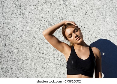 Outdoor Shot Of Beautiful Blonde Caucasian Woman Athlete Wearing Black Sportsbra Warming Up Neck Before Workout, Standing Against Concrete Wall Background With Copy Space For Your Promotional Content