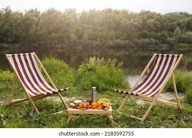 Outdoor Shot Of Beautiful Place For Picnic, Wooden Table With Food And Arm Chair For Relaxing Near River, Beautiful Nature, Resting, Camping, Recreation.