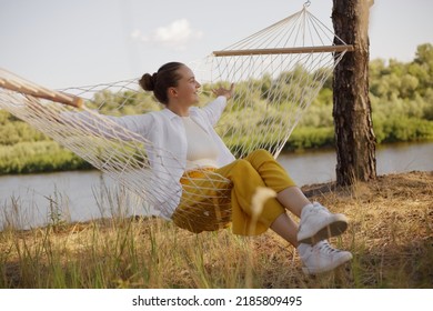 Outdoor shot of Caucasian young adult woman sitting on hammock by the water, looking away, resting, spreads hands, laughing happily, wearing white shirt and yellow trousers.