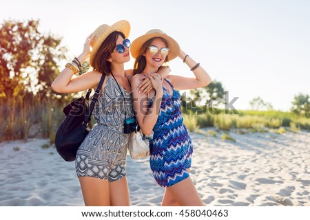 Outdoor shot of cheerful young female friends together on a beach at sunset. Two attractive women enjoying a holiday on the sea shore. Wearing trendy  summer outfit, backpacks , sunglasses. 