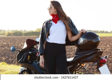 Outdoor Shot Of Confident Risky Young Woman Standing Near Her Motorbike, Putting Hand On Helmet, Wearing Red Bandana, White Shirt, Leather Jacket And Black Trousers. Motorcycle Tourism Concept.