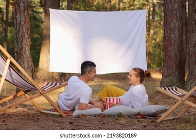 Outdoor Shot Of Couple Lying In The Forest With Overhead Projector With White Blank Screen, Eating Popcorn, Talking And Enjoying Their Leisure Time Together In The Nature.