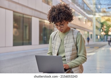 Outdoor shot of curly female student with curly hair dressed in casual clothes uses modern laptop prepares course work or new project poses in city. Female entrepreneur works remotely outside