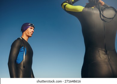 Outdoor Shot Of A Determined Male Triathlete Wearing Wetsuit Ready For Triathlon Competition With A Female. Athletic Swimmer Standing With His Swimming Gear On Against Sky.