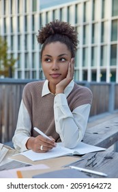 Outdoor Shot Of Female Student Prepares For Text Exam Writes Essay In Notebook Does Homework Puts Down Records To Daily Planner Sits Outside Dressed Neat Clothes Poses At Urban Setting Does Task