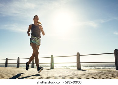 Outdoor shot of fitness young woman jogging along the beach. Female runner running outdoors on seaside promenade. - Powered by Shutterstock