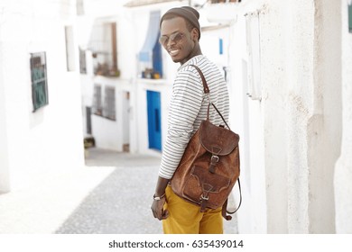 Outdoor Shot Of Handsome Happy Young African American Male Traveler With Leather Backpack Standing At Concrete Wall On Narrow Street While Sightseeing In Resort Town During His Summer Holidays