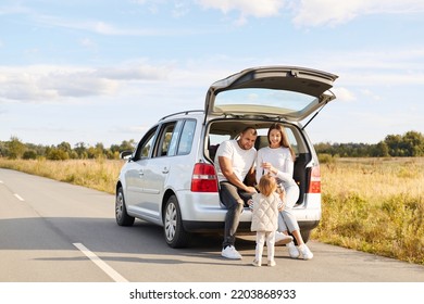 Outdoor Shot Of Happy Couple With Their Infant Daughter Sitting In Car Trunk And Drinking Hot Beverage, Coffee Or Tea From Thermos, Enjoying To Have Journey Together.