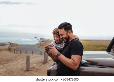 Outdoor Shot Of Happy Father And Son In Front Of The Car Using Mobile Phone And Laughing. Young Man And Little Boy Watching Something On Smart Phone And Enjoying While On Road Trip.