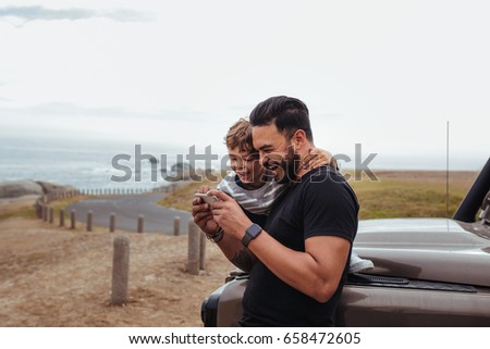 Outdoor shot of happy father and son in front of the car using mobile phone and laughing. Young man and little boy watching something on smart phone and enjoying while on road trip.
