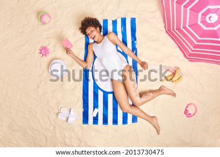 Outdoor shot of happy slim Afro American woman with curly hair dressed in white bikini holds passport and net bag of fruits spends vacation at seaside poses on striped towel enjoys summer holidays