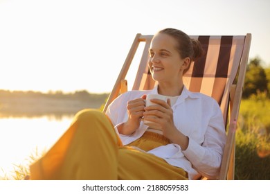 Outdoor Shot Of Happy Smiling Dark Haired Female Wearing White T Shirt Sitting In Folding Chair Near The River, Looking Away With Delighted Expression, Enjoying Coffee Or Tea.