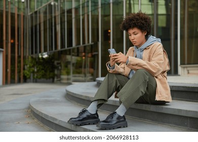 Outdoor Shot Of Serious Curly Haired Woma Dressed In Streetwear Uses Mobile Phone Browses Web Pages Sits On Steps Near Modern City Building Makes Post In Social Networks Communicates In Chat