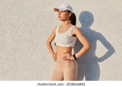 Outdoor shot of slim winsome female wearing sport white top, visor cap and leggins, standing against white wall, holding hands on hips, sporty woman, healthy lifestyle. - Powered by Shutterstock