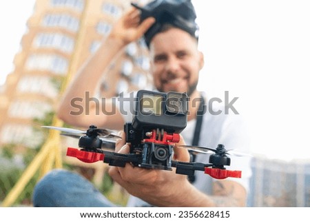 Outdoor shot of smiling drone pilot man holding fpv multicopter reaching to the camera. Focus is on hand with copter.