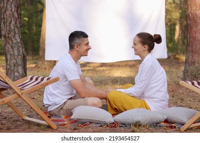 Outdoor Shot Of Smiling Satisfied Couple, Husband And Wife, Sitting In The Forest With Overhead Projector With Empty Screen On Background, Holding Hands And Talking.