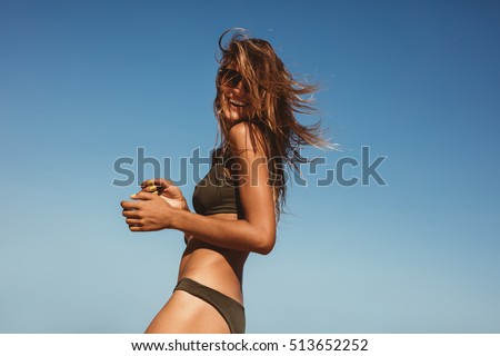 Outdoor shot of smiling young female model in bikini standing against blue sky. Woman having fun out on a summer day.