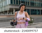 Outdoor shot of sporty fit woman poses with sport equipment checks time on smartwatch uses smartphone for tracking fitness results dressed in sportswear poses outdoors against urban background
